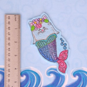 May include: Embroidered patch of a white cat with a flower crown, wearing a mermaid tail with a blue, green, and purple scale pattern. The tail has a pink fin. The patch is on a light blue background with blue waves.