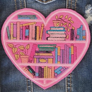 May include: A pink heart-shaped embroidered patch with a bookshelf design. The patch features colourful books, potted plants, and a detailed stitch pattern. The patch is likely for clothing or accessories.