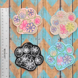 Peut inclure: Quatre patchs brodés en forme de patte d'animal, dans différentes couleurs : rose, beige, noir et bleu. Chaque patch présente un motif floral avec des points blancs.
