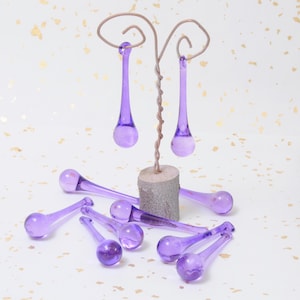 May include: A set of purple glass teardrop ornaments hanging from a wire stand. The ornaments are arranged in a cluster on a white background with gold flecks.