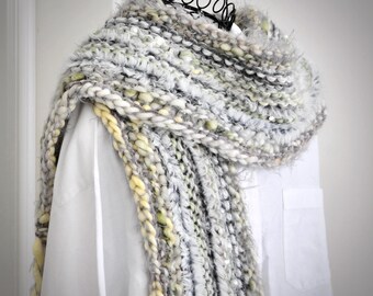May include: A chunky knit scarf featuring a blend of grey, white, and pale yellow yarn. The texture is varied, incorporating thick, fuzzy, and smooth strands with subtle metallic accents woven throughout the fabric, draped over a white shirt.