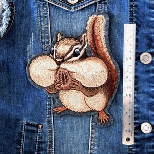 May include: An embroidered patch of a chipmunk with full cheeks holding a nut, positioned next to a metal ruler showing a height of approximately 5.5 inches. The patch features shades of brown, tan, and cream thread on a dark gray felt backing.