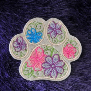 May include: A beige felt paw print with embroidered flowers in shades of pink, purple, blue and green.
