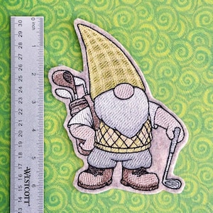 May include: Embroidered patch of a golf-playing gnome. The gnome wears a yellow hat, a gray beard, and a yellow and black vest. He carries a golf club and a bag of clubs. The patch is approximately 4 inches tall.