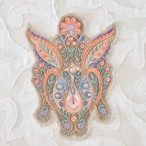 May include: An intricately embroidered patch in a symmetrical design, featuring a blend of peach, blue, and green hues. The patch has a beige border and is adorned with floral and paisley-like patterns, creating a detailed and ornate aesthetic.