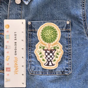 May include: Embroidered patch featuring a green topiary design in a black and white checkered vase. The patch has a cream-colored border and is set against a denim jacket pocket. The text "LOVE BEYOND Measure" is visible on a ruler.