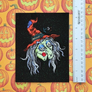 Könnte beinhalten: Ein schwarzer Filz-Patch mit glitzernder Oberfläche zeigt eine gestickte Abbildung einer Hexe mit grüner Haut, blauen Augen und einem roten Hut. Die Hexe hat langes, weißes Haar und trägt einen roten Hut mit schwarzem Rand. Der Hintergrund ist schwarz mit grünem Glitzer.