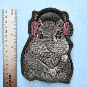 May include: Embroidered gray felt patch of a gray hamster wearing pink earmuffs and a black headband.