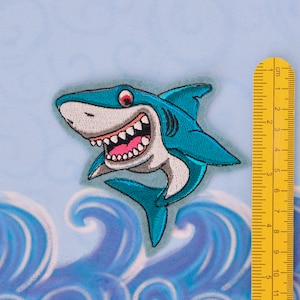 Può includere: Un toppa di squalo ricamata in blu e bianco, con un ghigno pieno di denti, che misura circa 6 cm di lunghezza. La toppa è posizionata su uno sfondo di onde blu e bianche.