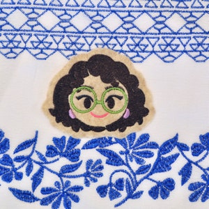 May include: Embroidered patch of a cartoon woman with brown hair, wearing green glasses and pink earrings. The patch is on a white background with blue embroidered flowers.