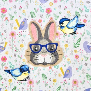 May include: A gray bunny with pink ears and blue glasses is surrounded by embroidered blue birds and flowers on a white background.