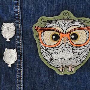 May include: A gray and white embroidered owl wearing orange-rimmed glasses. The owl is on a blue denim background.
