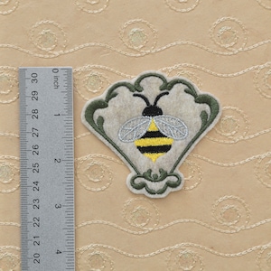 May include: A yellow and black embroidered bee with silver wings is centered on a beige felt applique. The applique is shaped like a heart with green embroidered vines around the edges.