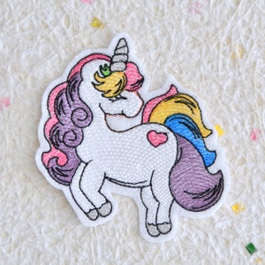 May include: Embroidered iron-on patch featuring a white unicorn with a pink, purple, and yellow mane and tail. The unicorn has a silver horn and a green eye. The unicorn has a pink heart on its side.