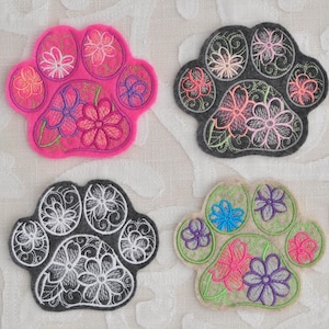 May include: Four embroidered felt paw prints with floral designs. The paw prints are in pink, gray, black, and beige.