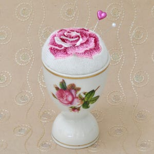May include: A white ceramic egg cup with a pink embroidered rose on top. The egg cup has a floral design and a gold trim.
