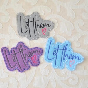 May include: Three felt patches in gray, purple, and blue, each with the phrase "Let them" in cursive script and a pink heart. The patches have a stitched border and are suitable for clothing or accessories.