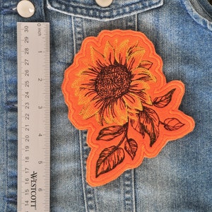 Puede incluir: Parche de fieltro naranja con un diseño de girasol bordado. El girasol tiene un contorno dorado y detalles negros. El tallo tiene hojas con contornos negros.