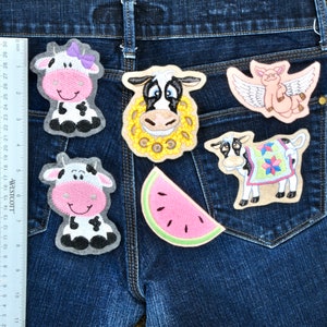 May include: A collection of embroidered patches featuring farm animals and a watermelon slice. The patches include a cow with a bow, a cow with a sunflower mane, a pig with wings, a cow with a blanket, and a watermelon slice. The patches are displayed on a blue denim background.