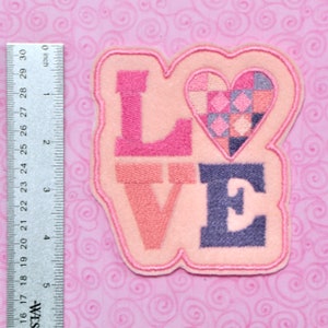 May include: Pink felt patch with the word "LOVE" embroidered in pink, peach, and blue thread. A patchwork heart is in the center of the patch.