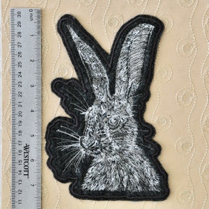 May include: A black and white embroidered patch of a rabbit's head with long ears. The patch is made of felt and has a black outline.