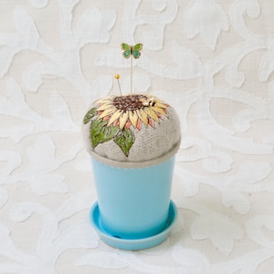 May include: A light blue ceramic pin cushion in the shape of a flower pot, with a sunflower design on top. A small butterfly pin and a gold pin are inserted into the cushion. The cushion is on a matching saucer.