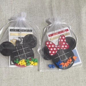 Op de afbeelding: Twee doorzichtige organzazakjes met elk een zwart Mickey Mouse-vormig Tic-Tac-Toe-spel. Eén heeft een rood-witte polkadot strik. Elk spel bevat kleurrijke stukken en een kleine kaart.