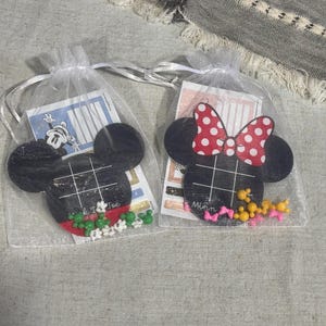 May include: Two white organza drawstring bags containing black wooden Mickey Mouse-shaped tic-tac-toe boards. One board features green and white glove-shaped game pieces, while the other includes yellow and pink bow-shaped pieces and a red polka-dot bow.