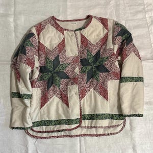 Könnte beinhalten: Gesteppte Jacke mit Patchwork-Design. Die Jacke hat eine cremefarbene Basis mit Stoffen in Rot, Grün und floralen Mustern. Das Design umfasst Sternmuster und grüne Blumenborte an Saum und Ärmeln. Sie hat eine Knopfleiste.