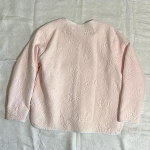 May include: A light pink quilted jacket with a floral pattern. The jacket has long sleeves and a rounded neckline. The fabric has a textured appearance, and the jacket is likely made of a soft material.