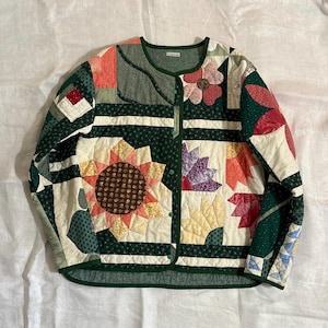 May include: A quilted jacket with a patchwork design featuring floral and geometric patterns. The jacket has a green trim and button closure. The design includes sunflowers, flowers, and various shapes in shades of green, red, yellow, and cream.