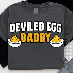 Può includere: T-shirt grigio scuro con la scritta "DEVILED EGG DADDY" in lettere bianche e gialle. Il design include due illustrazioni di uova farcite in ciotole bianche.