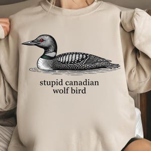 May include: Beige sweatshirt featuring a detailed illustration of a loon with the text "stupid canadian wolf bird" in black. The loon is depicted in black and white with a red eye, resting on the water.