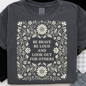 May include: Dark gray t-shirt with a floral design and the text "BE BRAVE BE LOUD AND LOOK OUT FOR OTHERS" in white. The design features flowers and leaves in a symmetrical pattern, creating a decorative border around the text.