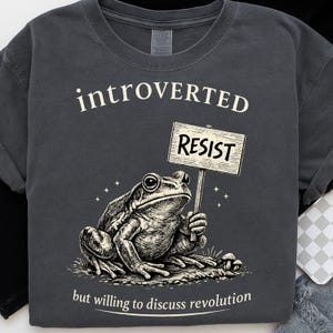 May include: Dark gray t-shirt with the word "introverted" above a frog illustration holding a "resist" sign. The text "but willing to discuss revolution" is below the frog. The design is in black and white.