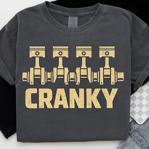 May include: Dark gray t-shirt featuring a distressed graphic of engine pistons and crankshafts. The word "CRANKY" is printed in a bold, yellow font below the engine illustration. A casual, comfortable tee.