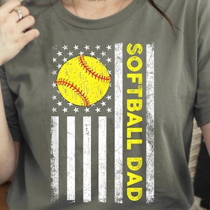 May include: Olive green t-shirt featuring a distressed American flag design with a yellow softball and the words "SOFTBALL DAD" in yellow. The shirt has a vintage, worn look.