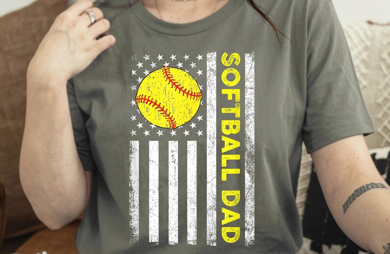 May include: Olive green t-shirt featuring a distressed American flag design with a yellow softball and the words "SOFTBALL DAD" in yellow. The shirt has a vintage, worn look.