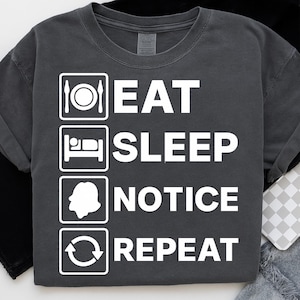 May include: Dark gray t-shirt with the words "EAT SLEEP NOTICE REPEAT" in white block letters. Each word is paired with a corresponding icon: plate and silverware, bed, a head, and a recycling symbol.