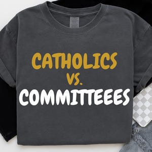 Op de afbeelding: Donkergrijs t-shirt met de tekst "CATHOLICS vs. COMMITTEES" in gouden en witte letters. Het shirt heeft een ronde hals en korte mouwen. De stof lijkt een zacht, comfortabel materiaal te zijn.