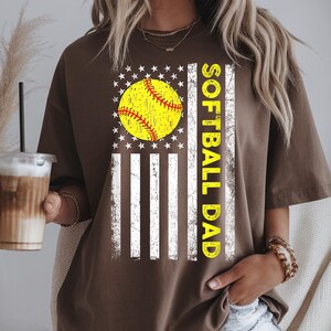 May include: A brown t-shirt featuring a softball design. The design includes a yellow softball with red stitching, set against a white and striped background with stars, and the words "SOFTBALL DAD" in yellow.