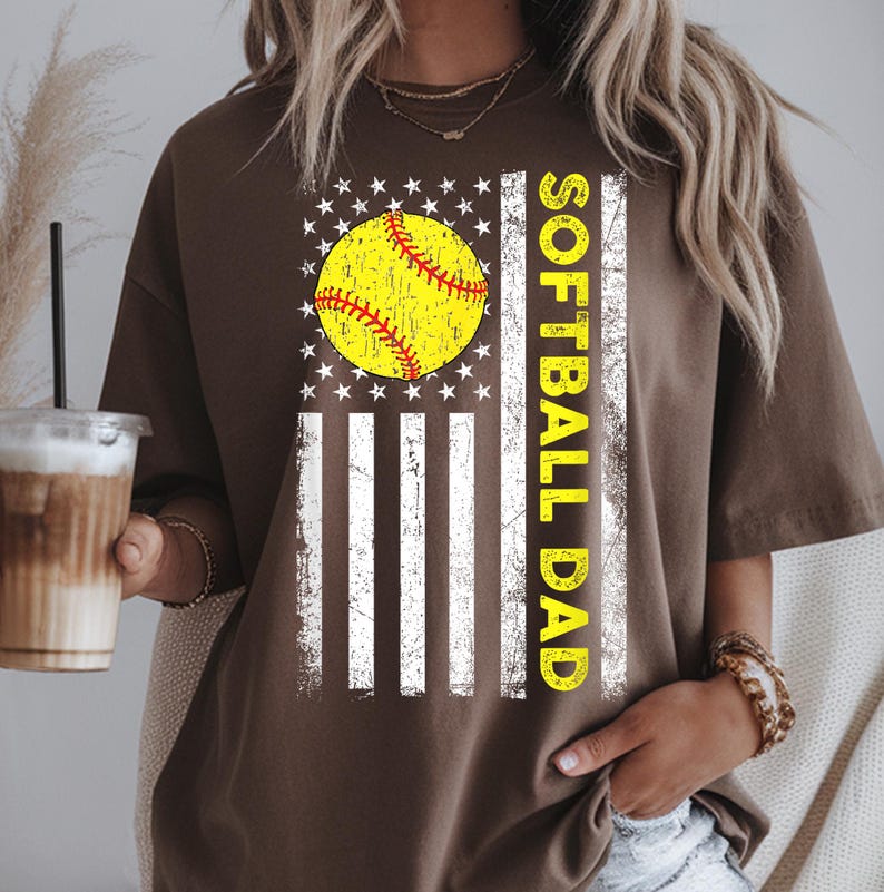 May include: A brown t-shirt featuring a softball design. The design includes a yellow softball with red stitching, set against a white and striped background with stars, and the words "SOFTBALL DAD" in yellow.
