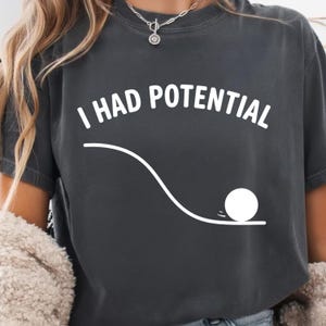 Pode incluir: Camiseta cinza escuro com o texto branco "I HAD POTENTIAL" acima de um gráfico branco de uma bola rolando por uma linha curva. A camiseta é de estilo casual.