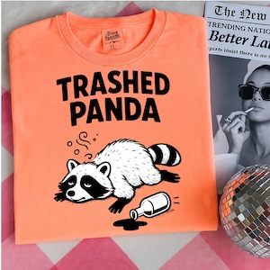 May include: An orange t-shirt featuring the text "TRASHED PANDA" in black. The shirt has a cartoon raccoon illustration, lying next to a spilled bottle. A newspaper and a disco ball are visible in the background.