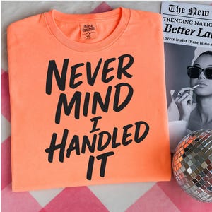 May include: A neon orange t-shirt with the phrase "NEVER MIND I HANDLED IT" in bold black letters. The shirt is folded and placed on a pink and white checkered surface. A newspaper and disco ball are in the background.