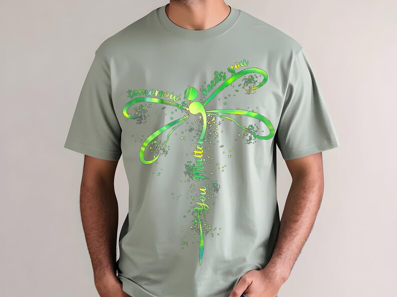 May include: A sage green t-shirt with a green and yellow dragonfly design. The dragonfly has the words "tomorrow needs you" written on its wings and "you matter" written on its body.