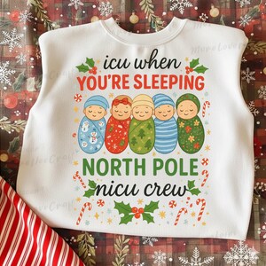 Puede incluir: Sudadera blanca de cuello redondo con un gráfico colorido. El gráfico presenta cinco bebés envueltos con diferentes patrones y colores, junto con el texto "icu when you're sleeping, North Pole nicu crew". Bastones de caramelo y acentos de acebo completan el diseño.