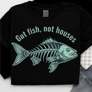 Könnte beinhalten: Schwarzes T-Shirt mit einem teal-farbenen Fischskelett-Design und dem Text "Gut fish, not houses". Die Grafik zeigt detailliert die Knochen, Flossen und den Kopf des Fisches. Text und Grafik sind in passendem Teal gehalten.