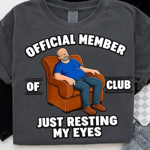 Könnte beinhalten: Dunkelgraues T-Shirt mit einer Cartoon-Illustration eines Mannes in einem braunen Sessel. Der Text lautet "OFFICIAL MEMBER OF CLUB" über dem Bild und "JUST RESTING MY EYES" darunter.