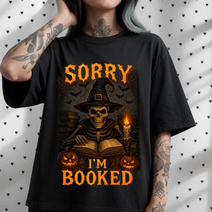 Peut inclure: T-shirt noir avec un motif sur le thème d'Halloween. Le motif représente un squelette portant un chapeau de sorcière, lisant un livre, avec des citrouilles et des chauves-souris. Le texte dit "SORRY I'M BOOKED".